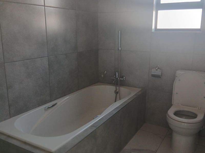 A modern bathroom featuring grey tiled walls, a built-in bathtub with shower mixer, and a standard toilet.