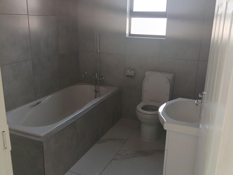 A modern, neutral-toned bathroom featuring a built-in bathtub, a white vanity basin, and a toilet, finished with contemporary wall and floor tiling.