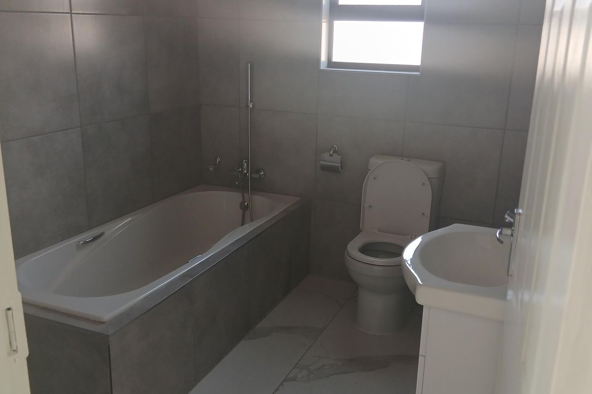 A modern, neutral-toned bathroom featuring a built-in bathtub, a white vanity basin, and a toilet, finished with contemporary wall and floor tiling.
