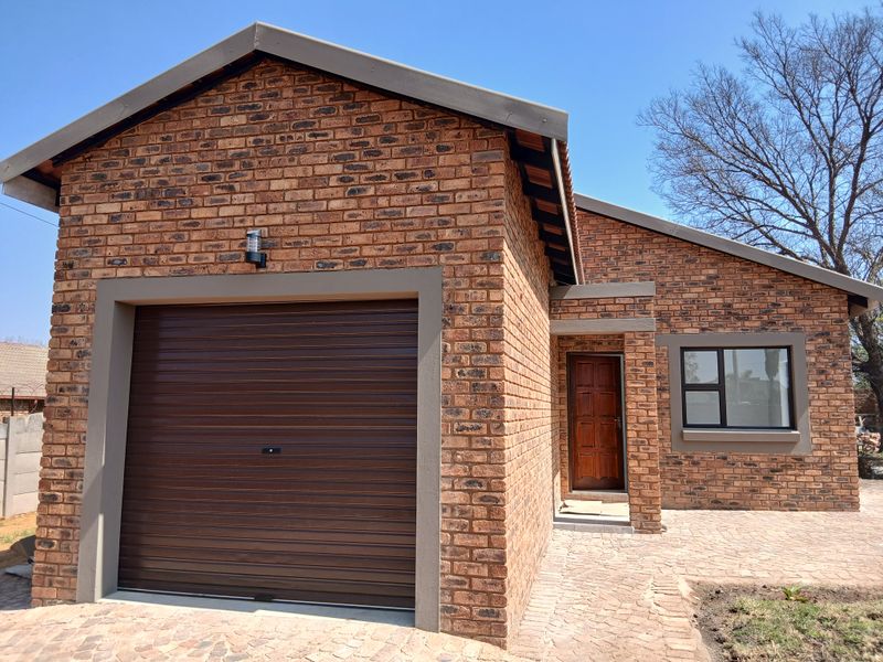A well-maintained face brick house exterior featuring a single roller shutter garage, timber front door, and paved driveway.