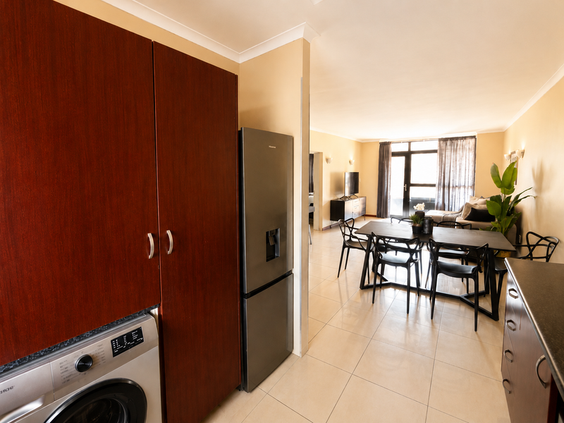 Open-plan living and dining area featuring tiled floors, built-in kitchen cabinetry with integrated laundry, and access to a balcony through sliding glass doors.