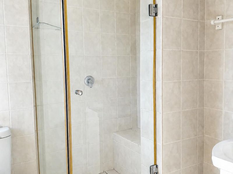 A functional bathroom featuring a glass-enclosed shower with a built-in bench, pedestal sink, and wall-mounted toilet, finished with neutral wall and floor tiles.
