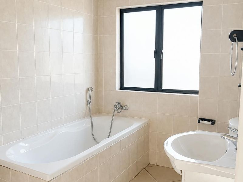 A clean, neutral-toned bathroom featuring a built-in bathtub, pedestal basin, and tiled finishes throughout, offering a functional and well-maintained space.