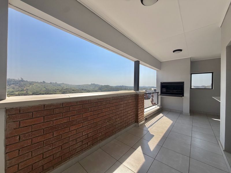 Spacious covered balcony featuring tiled flooring, a built-in braai, and a brick balustrade with expansive views of the surrounding landscape.