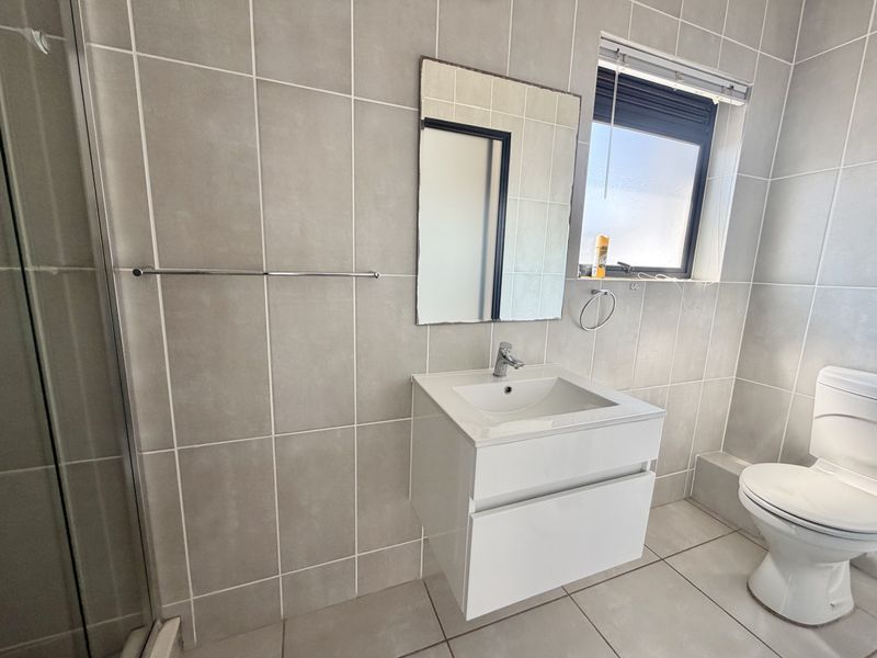 A modern, neutral-toned bathroom featuring a wall-mounted vanity, tiled walls and flooring, a shower enclosure, and a standard toilet.