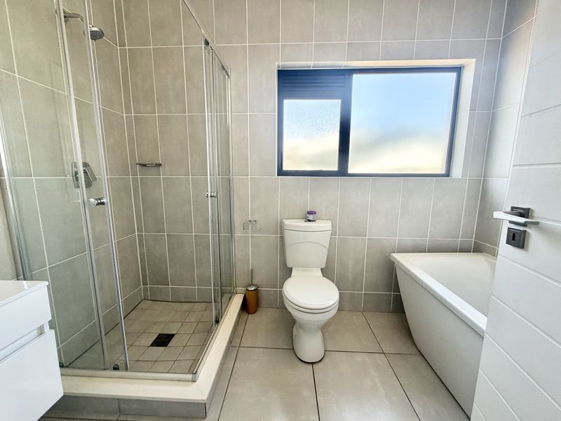 Modern bathroom featuring a glass-enclosed shower, a separate bathtub, a toilet, and neutral floor-to-ceiling tiling with a frosted window for natural light.