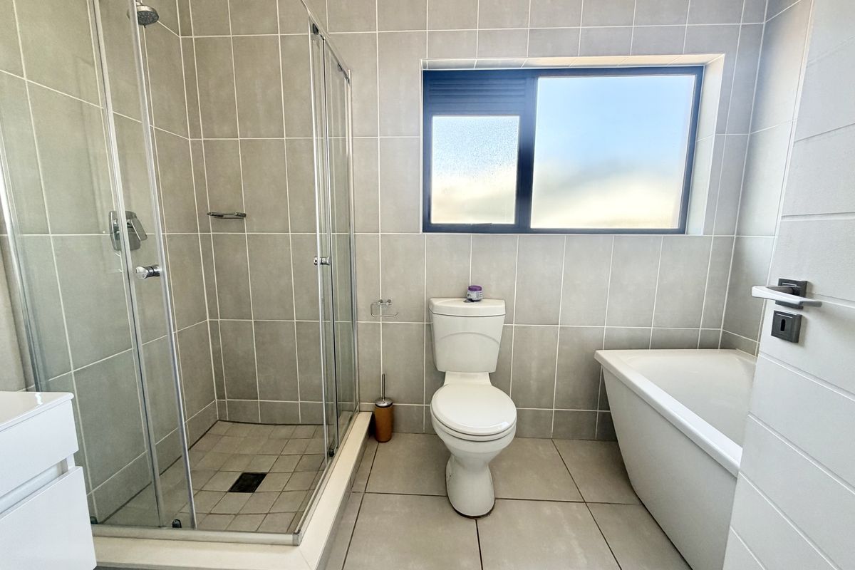 Modern bathroom featuring a glass-enclosed shower, a separate bathtub, a toilet, and neutral floor-to-ceiling tiling with a frosted window for natural light.