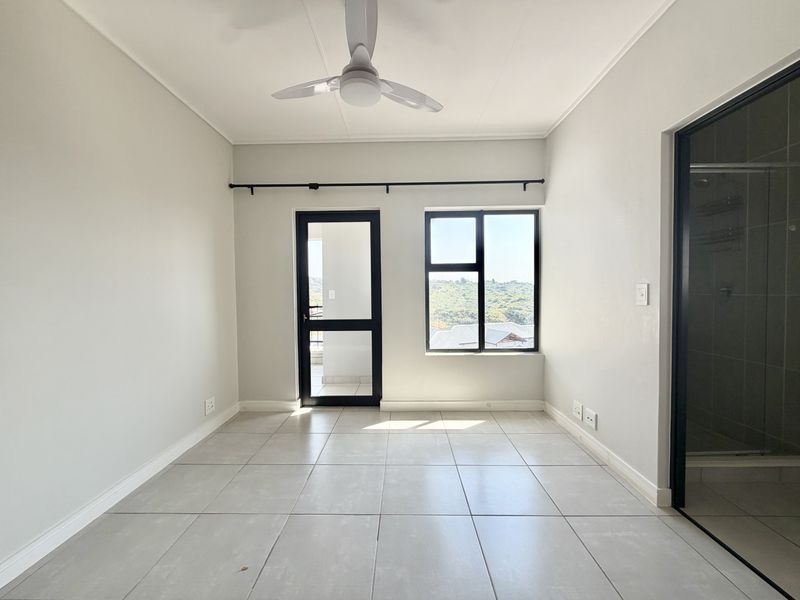 A bright, empty bedroom featuring neutral tiled flooring, a ceiling fan, and direct access to an outdoor balcony through an aluminium door.