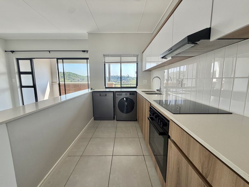 Modern kitchen featuring sleek wood-finish cabinetry, stone countertops, integrated appliances, and tiled flooring, with an open-plan layout and views of the surrounding landscape.