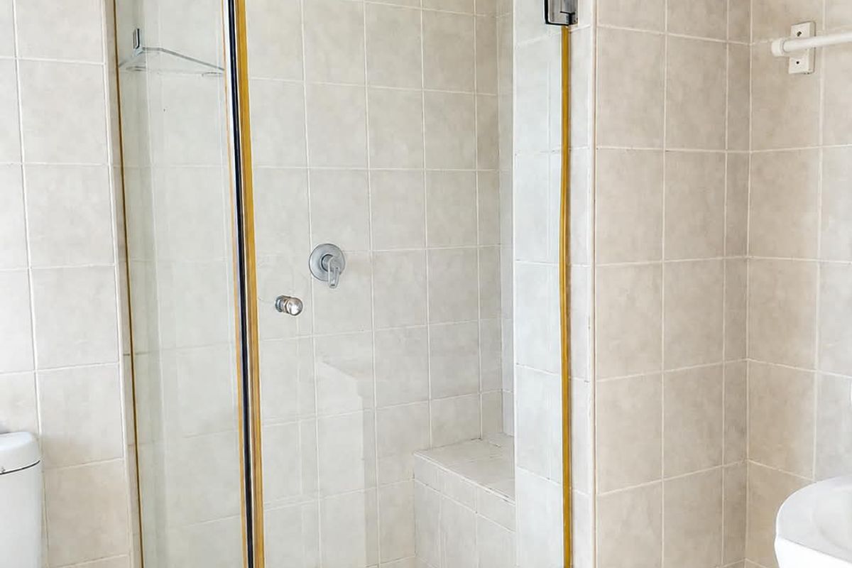 A functional bathroom featuring a glass-enclosed shower with a built-in bench, pedestal sink, and wall-mounted toilet, finished with neutral wall and floor tiles.