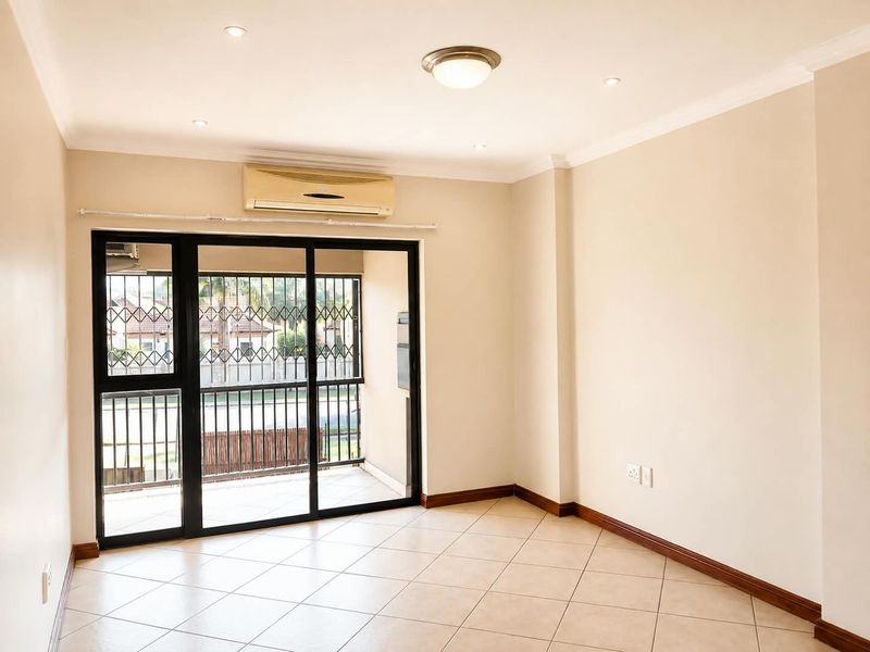 A bright, empty living room featuring tiled floors, recessed lighting, and a sliding glass door leading to a balcony with security gates.