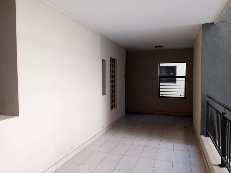 A spacious, covered balcony featuring neutral tiled flooring, a metal safety balustrade, and a decorative ventilation screen, offering a clean and functional outdoor area.