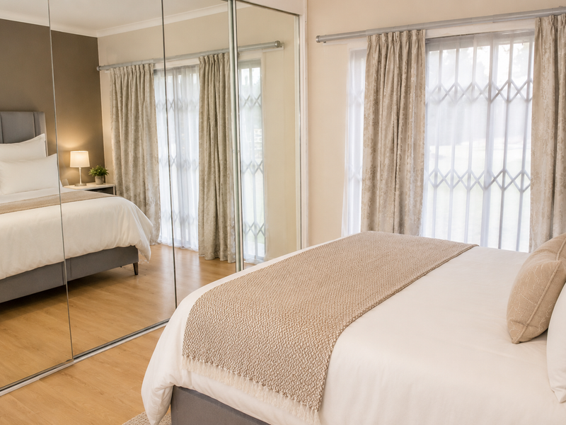 A well-maintained bedroom featuring light laminate flooring, large mirrored sliding wardrobe doors, and security gates on the windows for added peace of mind.