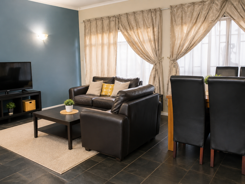 An open-plan living and dining area featuring dark tiled flooring, a blue accent wall, and large windows with neutral curtains.