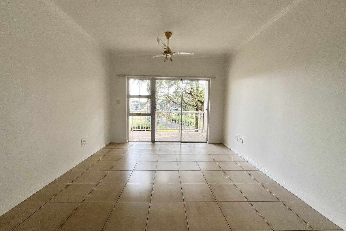 A bright, empty living room featuring neutral tiled flooring, a ceiling fan, and sliding glass doors that open onto a balcony.