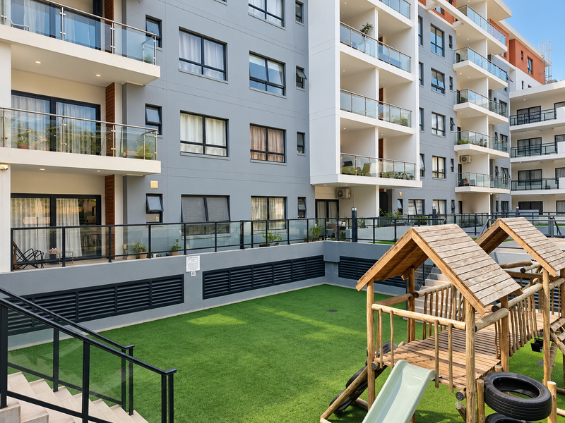 A modern apartment complex exterior featuring private balconies with glass balustrades overlooking a communal courtyard with an artificial lawn and wooden playground equipment.