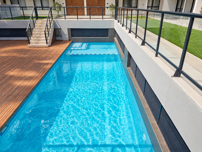 A modern swimming pool area featuring timber decking, glass balustrades, and a staircase leading to an elevated garden space.