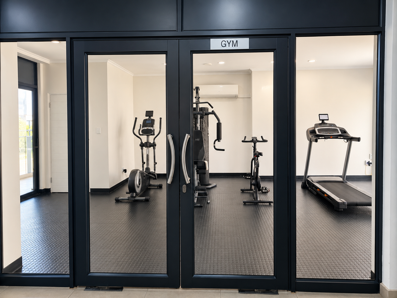 A well-maintained residential gym featuring durable rubber flooring, recessed lighting, and a split-system air conditioner, accessed through modern glass-paneled double doors.
