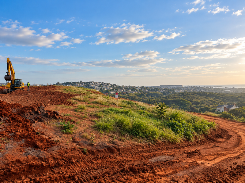 A vacant residential plot undergoing earthworks with an excavator on site, offering elevated views of the surrounding neighborhood and landscape.