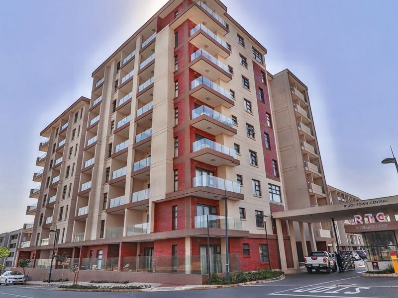 A modern multi-story apartment complex featuring a brick facade, glass balconies, and a secure entrance with a covered canopy and boom gate.