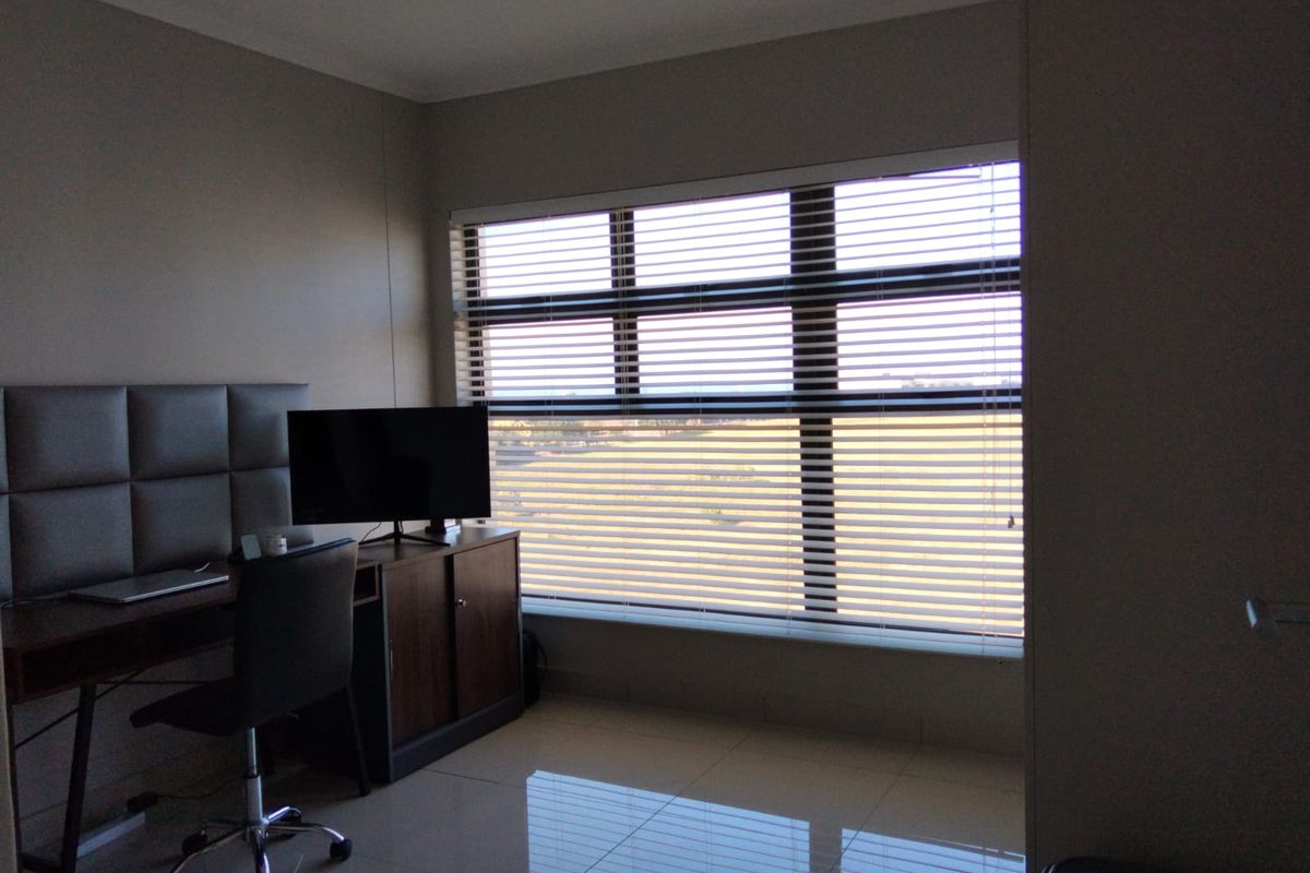 A bedroom featuring tiled flooring, an upholstered headboard, and large windows fitted with venetian blinds, offering natural light and an exterior view.