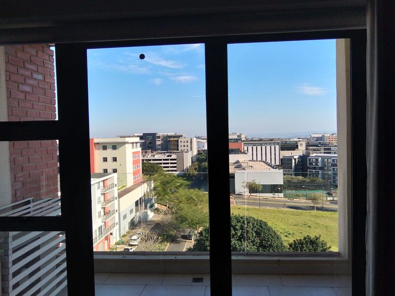 A clear urban view from a balcony featuring a glass balustrade and tiled flooring, overlooking surrounding apartment buildings and green space.