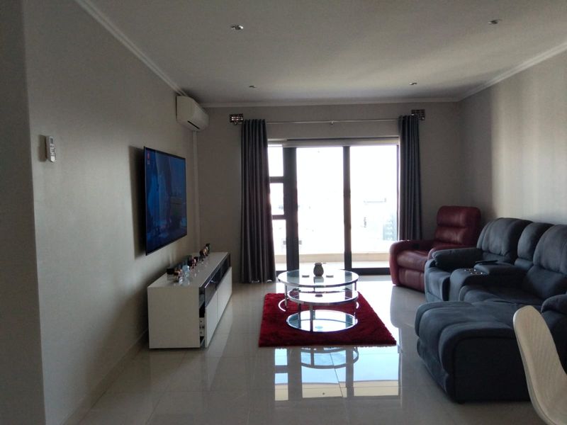 A well-maintained living room featuring tiled flooring, recessed downlighting, a wall-mounted air conditioning unit, and large sliding glass doors leading to a balcony.