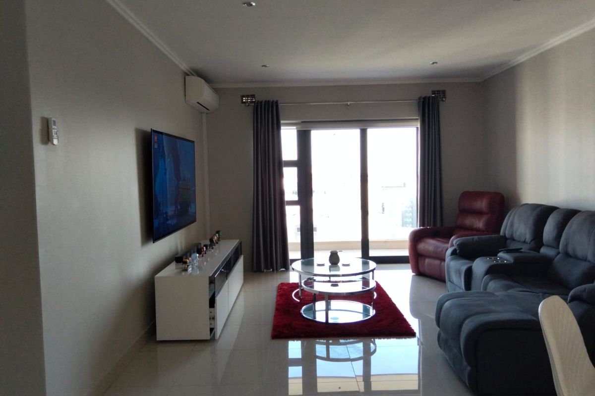 A well-maintained living room featuring tiled flooring, recessed downlighting, a wall-mounted air conditioning unit, and large sliding glass doors leading to a balcony.