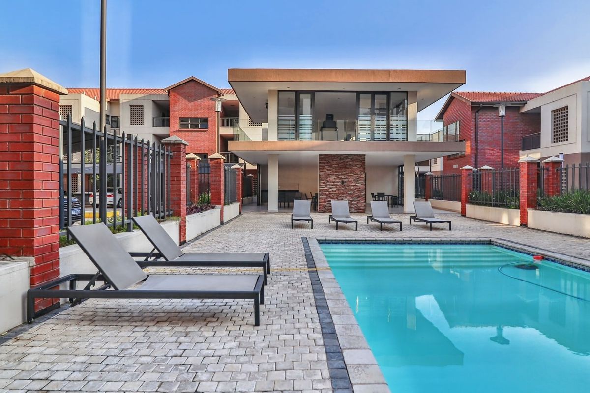 A well-maintained communal pool area featuring a paved deck, stone feature wall, and a covered patio structure within a brick-walled apartment complex.