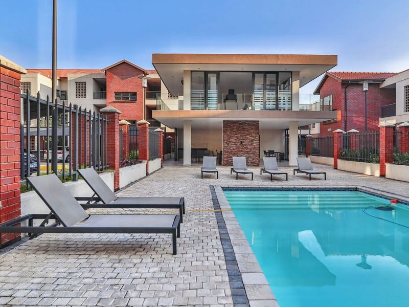 A well-maintained communal pool area featuring a paved deck, stone feature wall, and a covered patio structure within a brick-walled apartment complex.