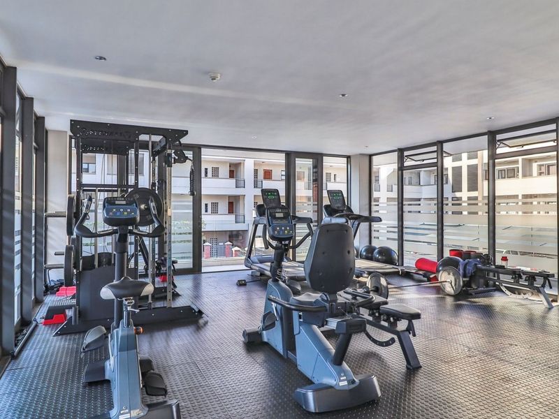 A well-lit, modern apartment gym featuring rubberized flooring and extensive floor-to-ceiling windows providing natural light and views of the surrounding complex.