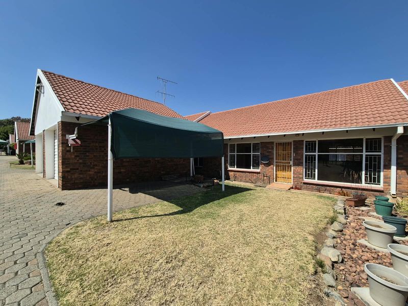 Rear view of a townhouse featuring a brick facade, tiled roof, paved driveway, and a small garden area with a shade sail.