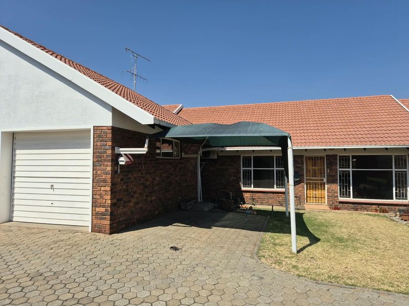 Front exterior view of a townhouse featuring a brick facade, tiled roof, paved driveway, single garage, and a shade cloth carport.