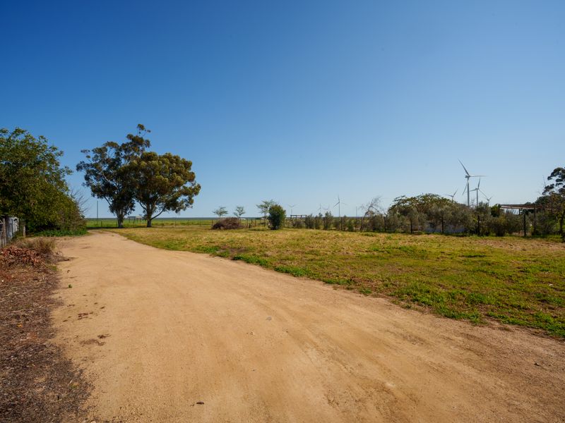 A view of a vacant residential plot featuring a dirt access road, established trees, and perimeter fencing under a clear blue sky.