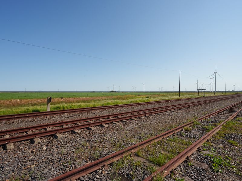 A wide view of the surrounding landscape featuring railway tracks, open fields, and distant wind turbines under a clear blue sky.