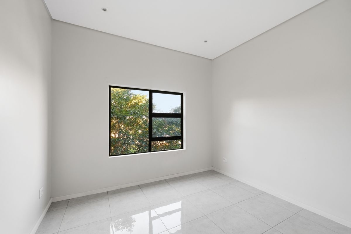 A bright, empty bedroom featuring neutral walls, modern tiled flooring, and a black aluminium window frame with a view of mature trees.