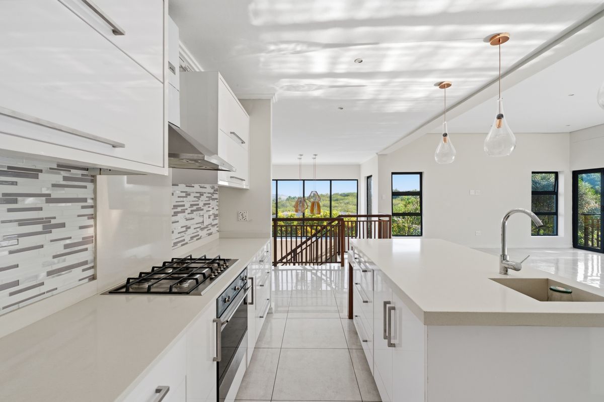 Modern kitchen featuring white cabinetry, stone countertops, a central island with sink, gas hob, and integrated oven, overlooking an open-plan living area.
