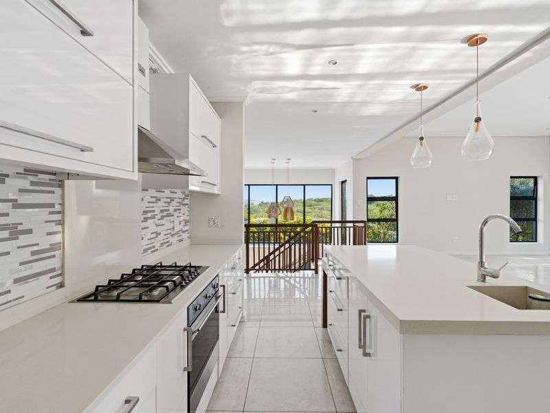 Modern kitchen featuring white cabinetry, stone countertops, a central island with sink, gas hob, and integrated oven, overlooking an open-plan living area.