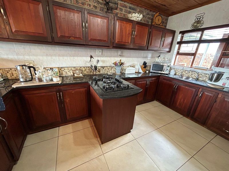 A functional kitchen featuring dark wood cabinetry, granite countertops, a gas hob, and a distinctive exposed brick wall with timber ceiling accents.