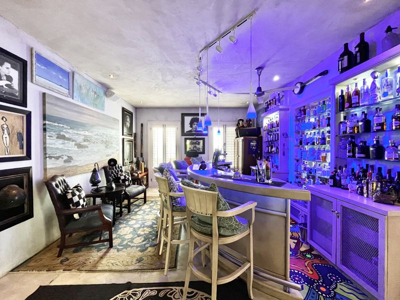 A well-appointed home bar area featuring a built-in counter, extensive illuminated shelving, track lighting, and shutter blinds, creating a vibrant entertainment space.