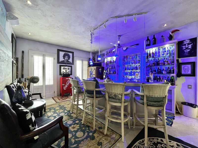 A dedicated home bar area featuring a built-in counter, extensive backlit shelving for display, track lighting, and direct access through french doors.