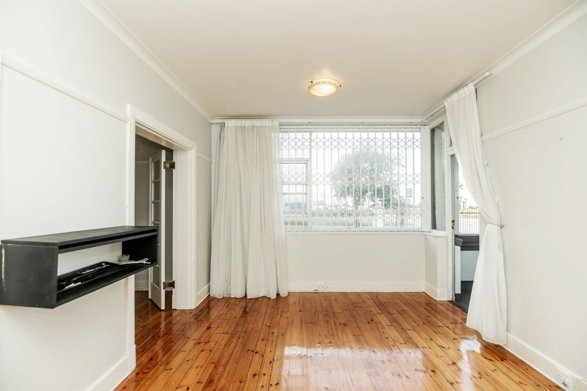 A bright, empty living room featuring polished hardwood flooring, coving, and a large window fitted with a security gate.