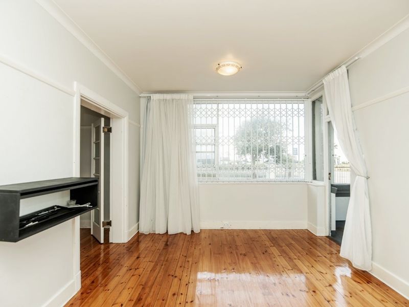 A bright, empty living room featuring polished hardwood flooring, coving, and a large window fitted with a security gate.