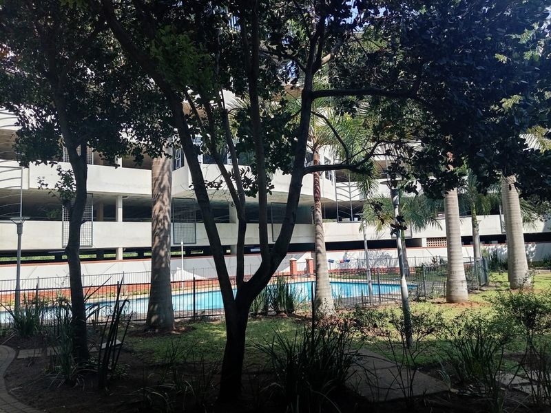 Communal swimming pool area featuring landscaped gardens and metal perimeter fencing, situated adjacent to a multi-level apartment parking structure.