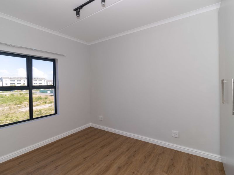 A bright, empty bedroom featuring modern laminate flooring, a built-in wardrobe, and a black-framed window with a view of the surrounding area.