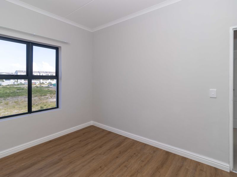 A bright, empty bedroom featuring modern laminate flooring, neutral walls, and a large aluminium-framed window with a view of the surrounding area.