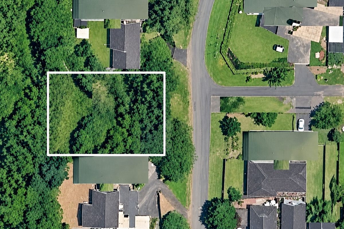 An aerial view of a residential neighborhood showing a vacant plot of land highlighted within a white border, surrounded by established homes and trees.