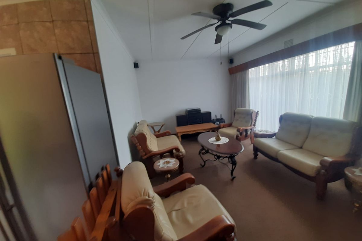 A living room featuring a ceiling fan, carpeted flooring, and a large window with security bars, offering a dated but functional living space.