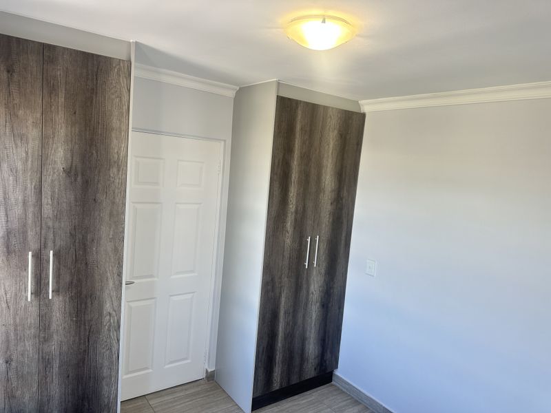 A clean, empty bedroom featuring modern wood-finish built-in cupboards, neutral walls, and tiled flooring with classic ceiling cornice detailing.