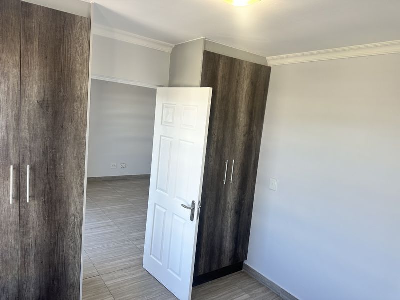 A clean, empty bedroom featuring modern wood-finish built-in cupboards, neutral walls, and tiled flooring with a view into an adjacent room.